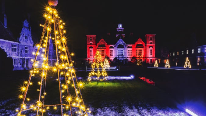 Image shows Christmas tree sculptures wound with fairy lights in front of the illuminated hall at Blickling